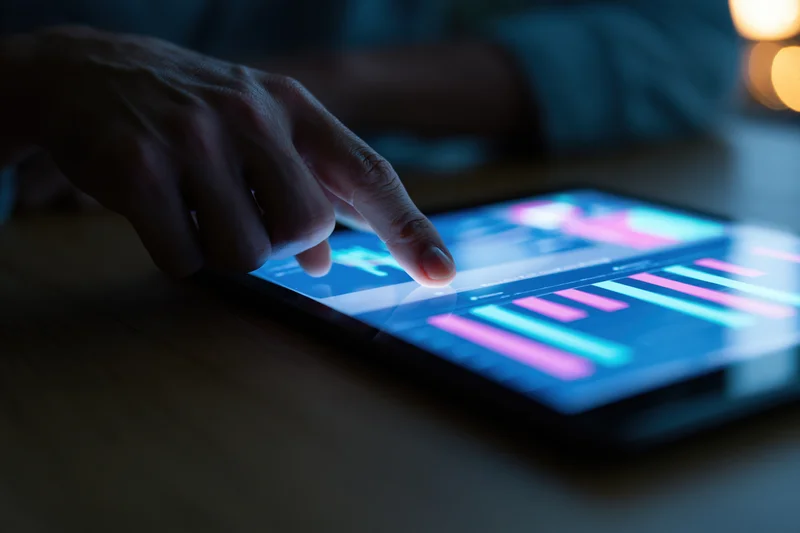 Hand tapping a glowing tablet screen displaying colorful data charts on a wooden surface at night