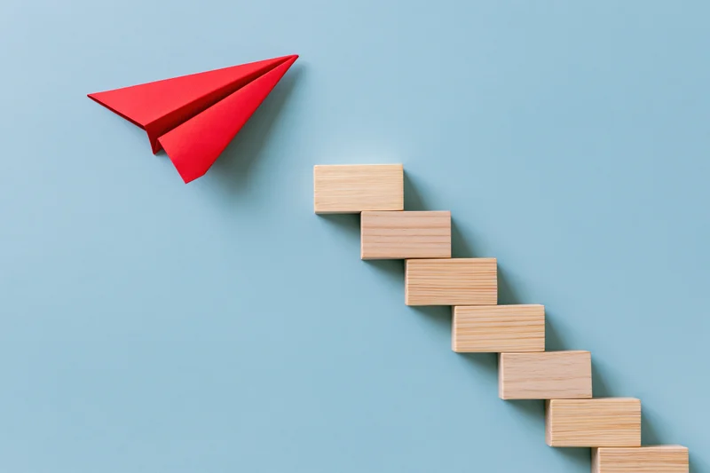 Red paper airplane flying upward beside a wooden block staircase on a blue background
