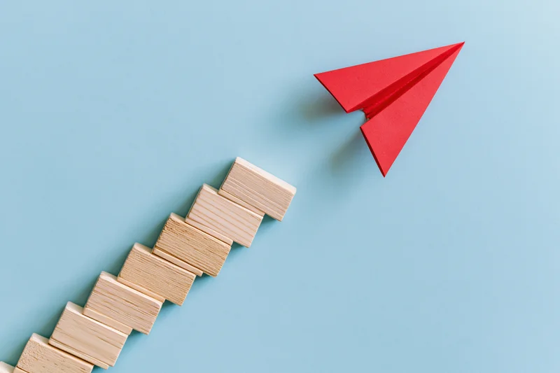Red origami paper airplane flying upward alongside a staircase of wooden blocks on blue background
