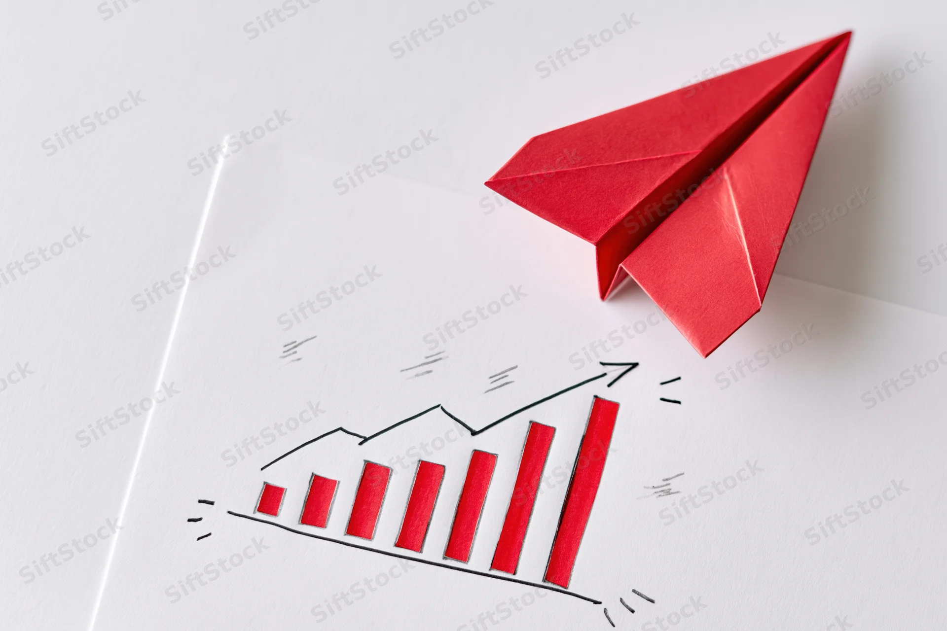 Red origami paper airplane resting on a white surface above a hand-drawn bar chart showing upward growth