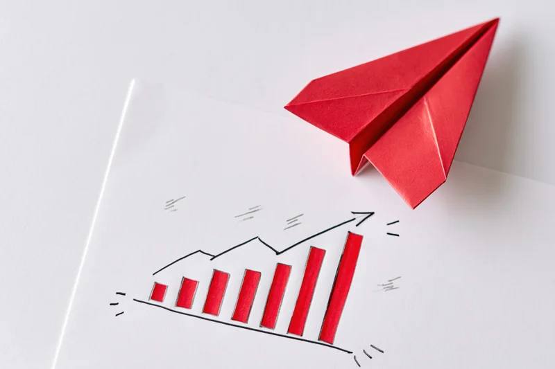 Red origami paper airplane resting on a white surface above a hand-drawn bar chart showing upward growth