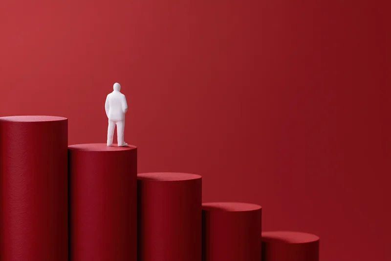 White figurine standing on top of red cylindrical columns arranged as a rising bar chart
