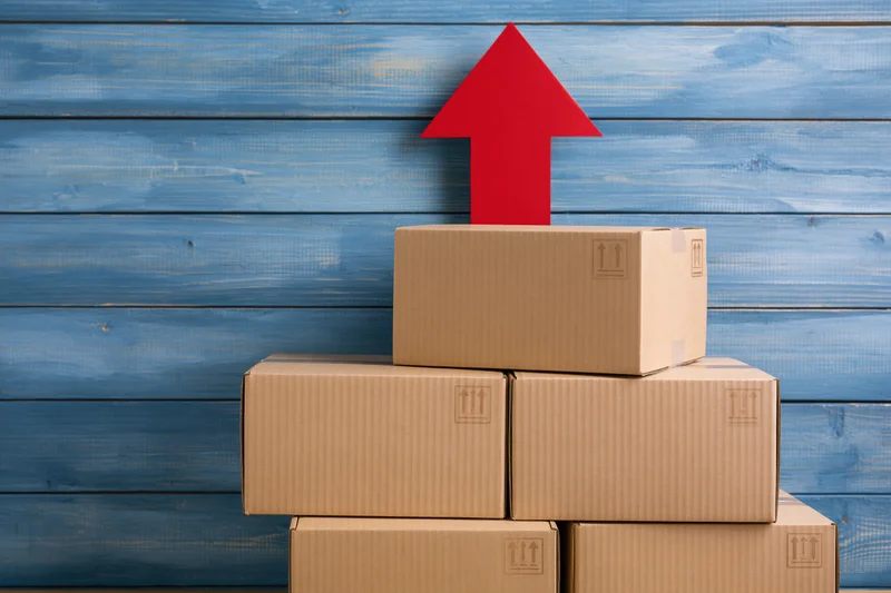 Stack of brown cardboard shipping boxes piled against blue wooden wall with red arrow pointing upward