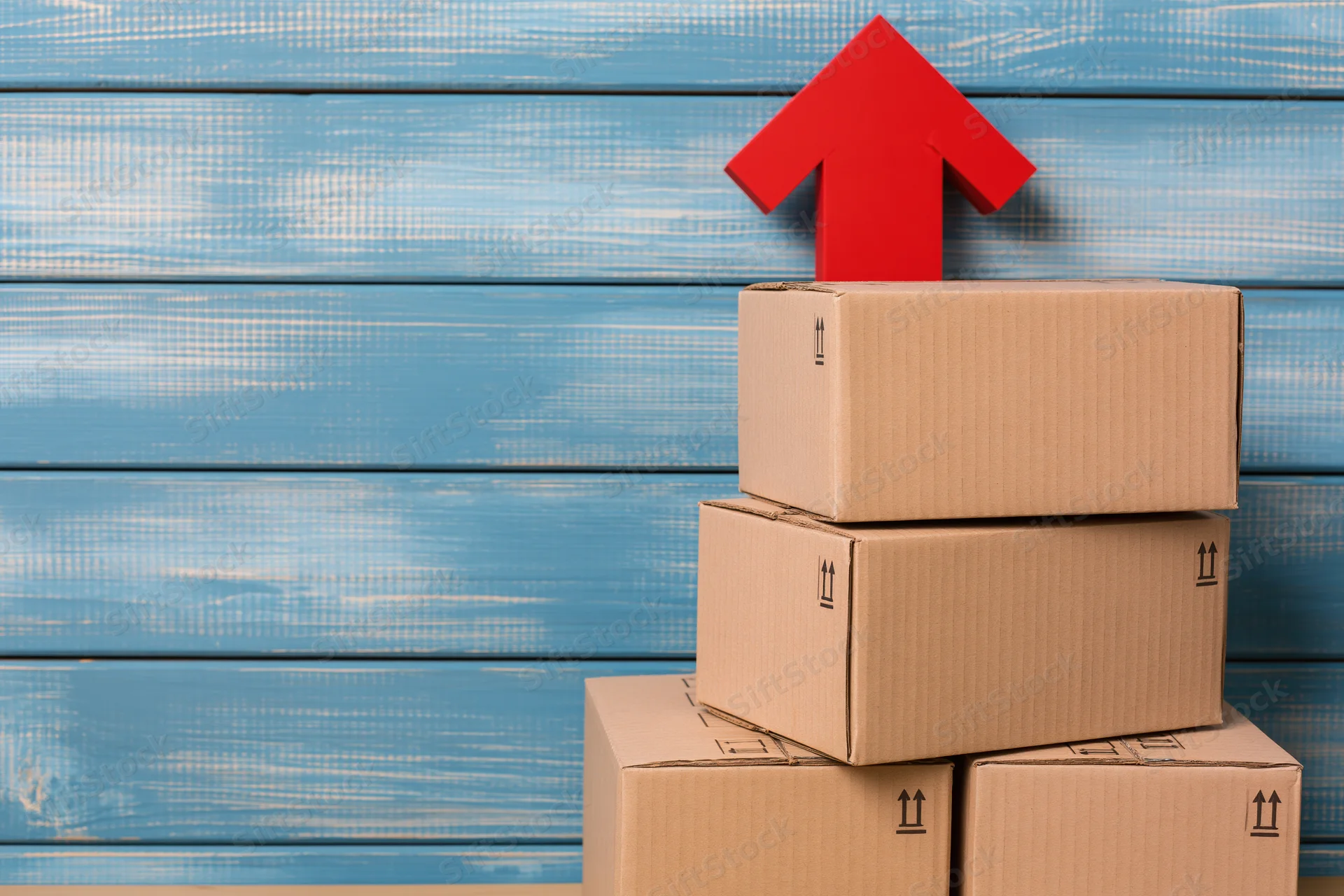 Stack of brown cardboard shipping boxes with red arrow pointing up against blue wooden background