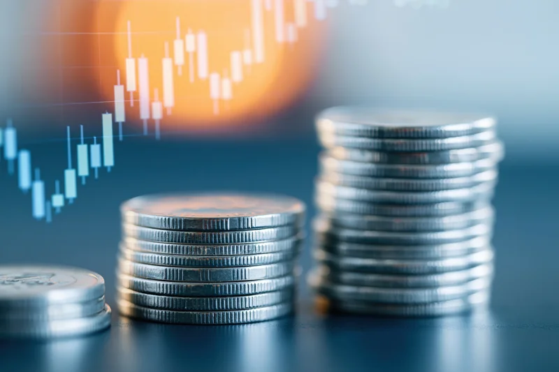 Stacks of silver coins on a dark surface with a digital stock chart in the background