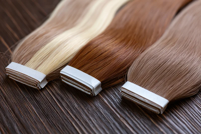Three bundles of human hair extensions in ash blonde and warm auburn tones arranged on a dark wooden surface