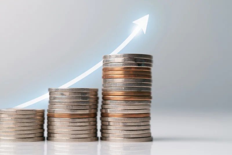 Three stacks of coins increasing in height with a glowing arrow rising behind them on a light surface