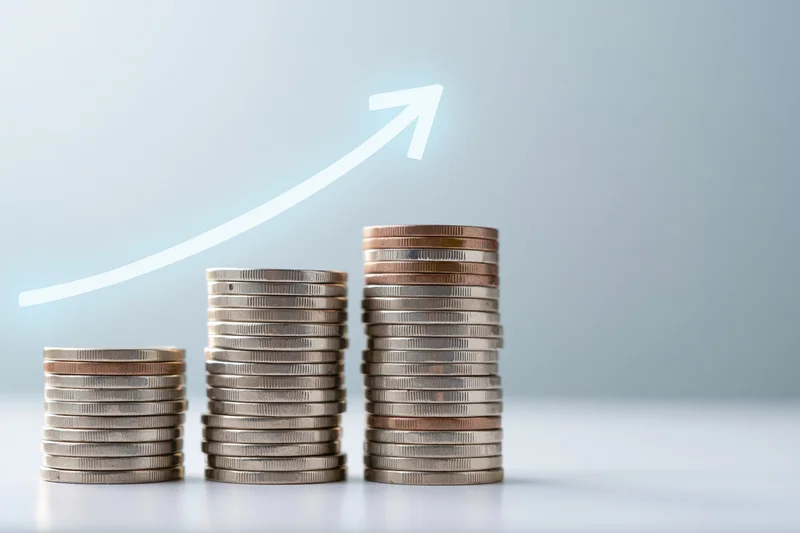 Three stacks of coins increasing in height with a glowing arrow pointing upward on a light background