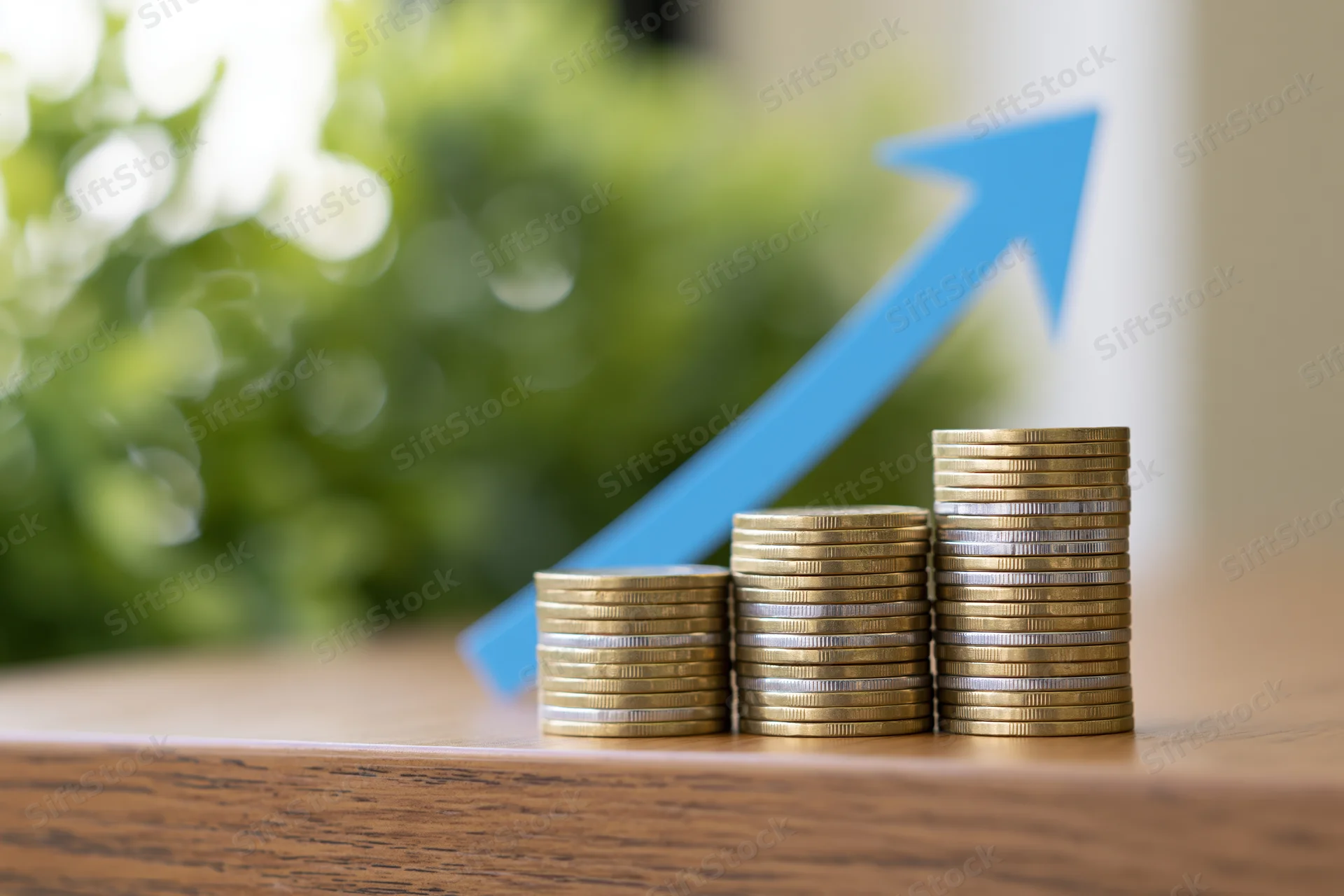 Three stacks of coins arranged in ascending height on a wooden table with a blue arrow pointing upward in the background