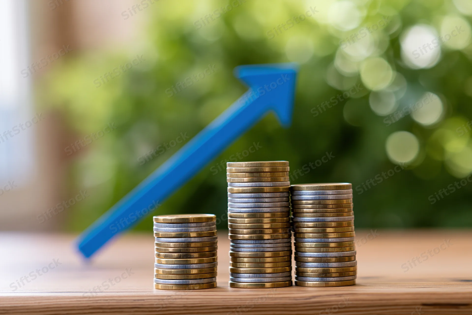Three stacks of coins increasing in height with a blue arrow pointing upward on a wooden table