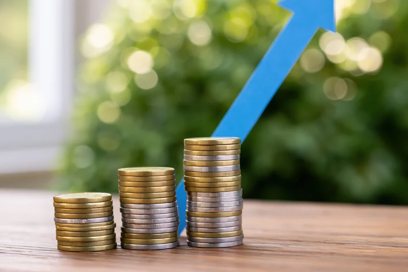 Three stacks of gold and silver coins arranged in ascending height on a wooden table with a blue upward arrow symbolizing growth