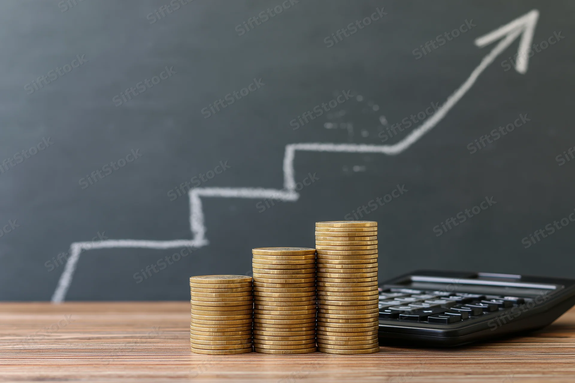 Three stacks of gold coins increasing in height on a wooden desk beside a calculator with a chalk-drawn upward arrow on a blackboard behind