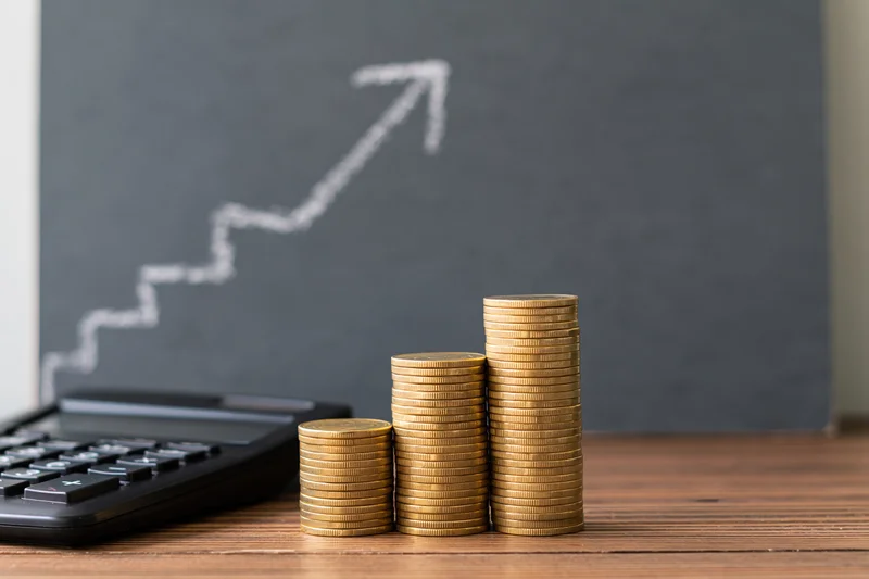 Three stacks of gold coins rising in height on a wooden desk beside a calculator with a chalk-drawn upward arrow on a blackboard behind them