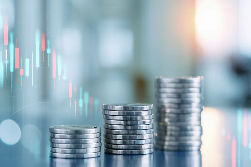 Three stacks of silver coins arranged in ascending height on a reflective surface with digital stock market charts overlaid in the background