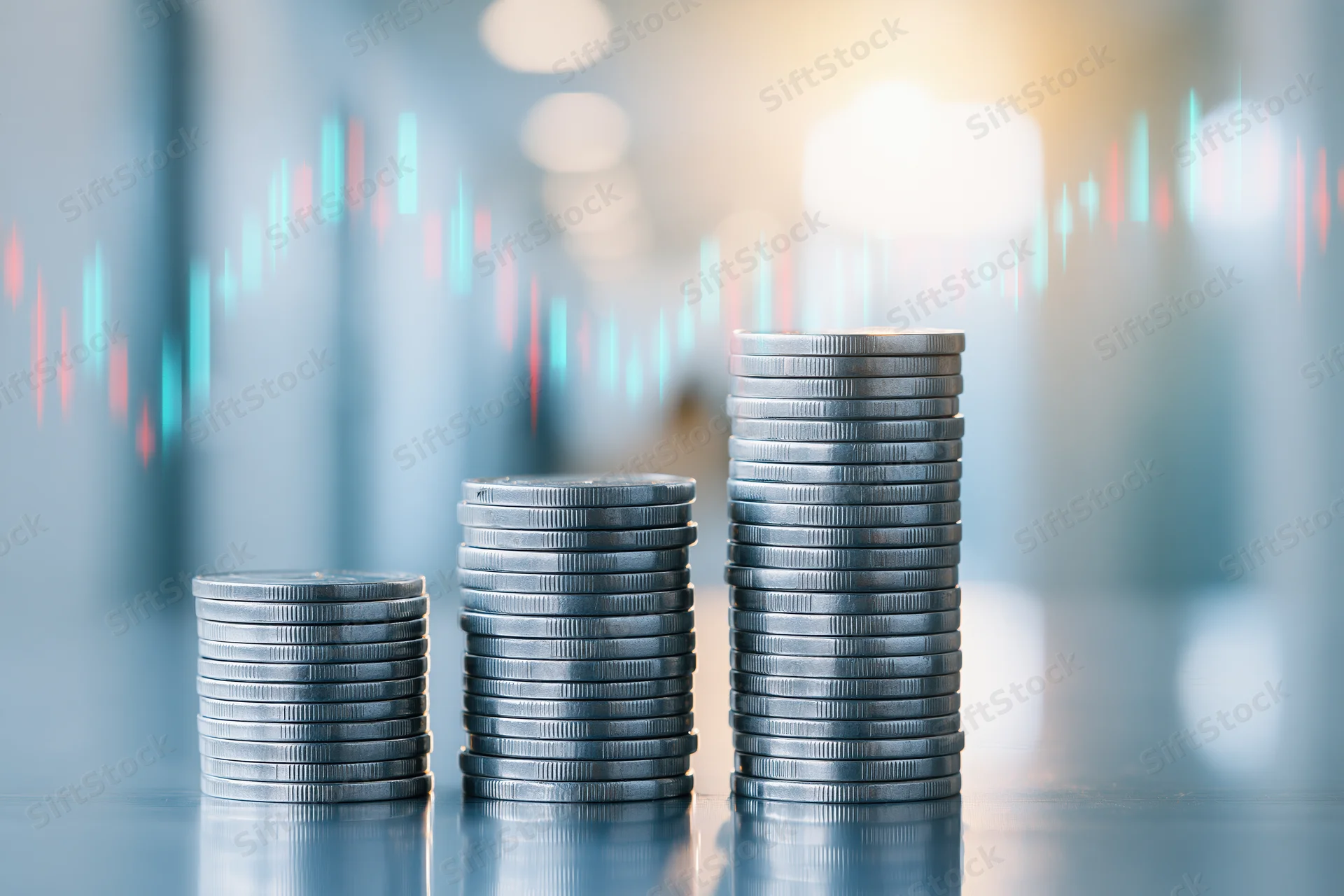 Three stacks of silver coins arranged in ascending height on a reflective surface with financial chart in background