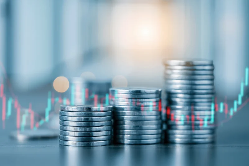 Three stacks of silver coins arranged in ascending height on a dark surface with overlaid stock market candlestick chart showing upward trend