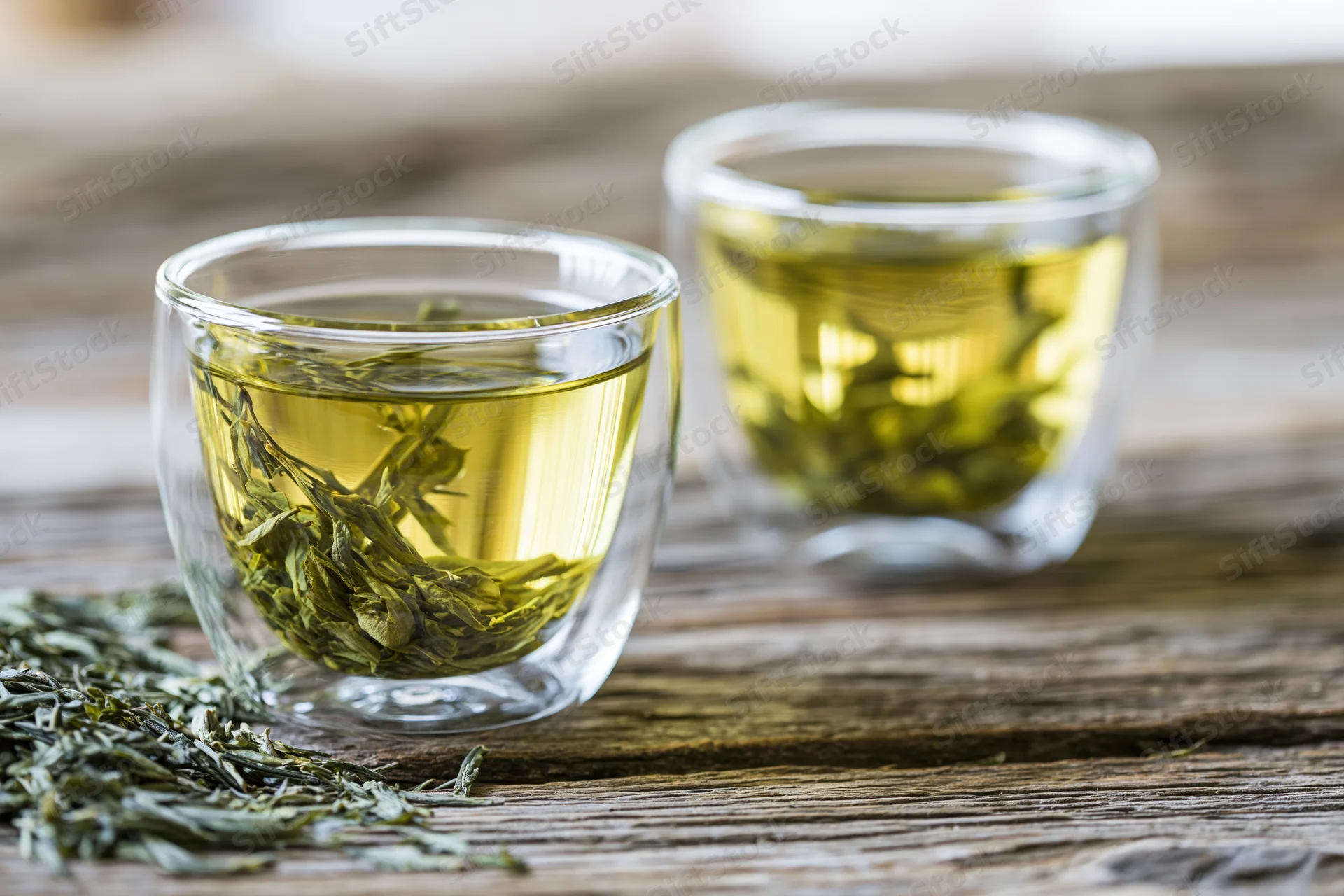 Two glass cups filled with brewed green tea and loose leaves on a wooden table