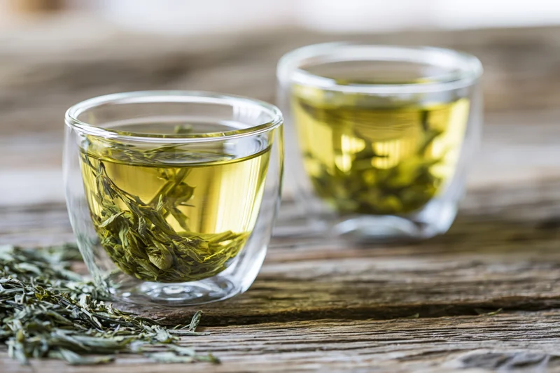 Two glass cups filled with brewed green tea and loose leaves on a wooden table