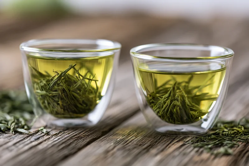 Two glass cups filled with brewed green tea and floating leaves on a wooden surface