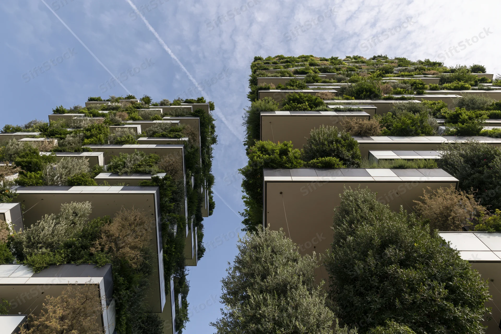 Two tall residential towers covered in greenery and trees, viewed from below against a blue sky