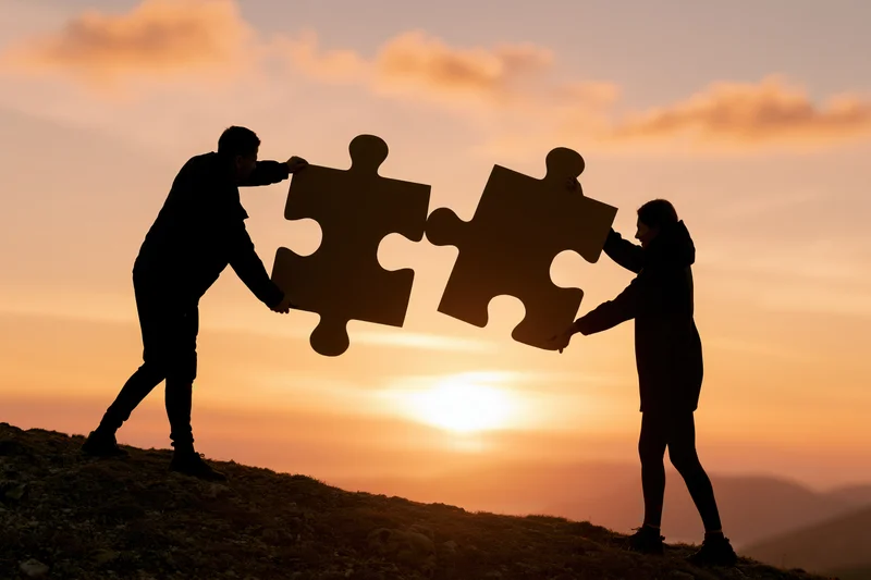 Two people connecting large puzzle pieces on a hill at sunset