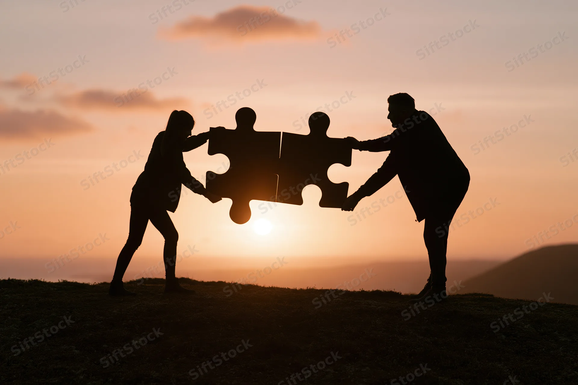 Two silhouetted figures on a hillside joining puzzle pieces at sunset