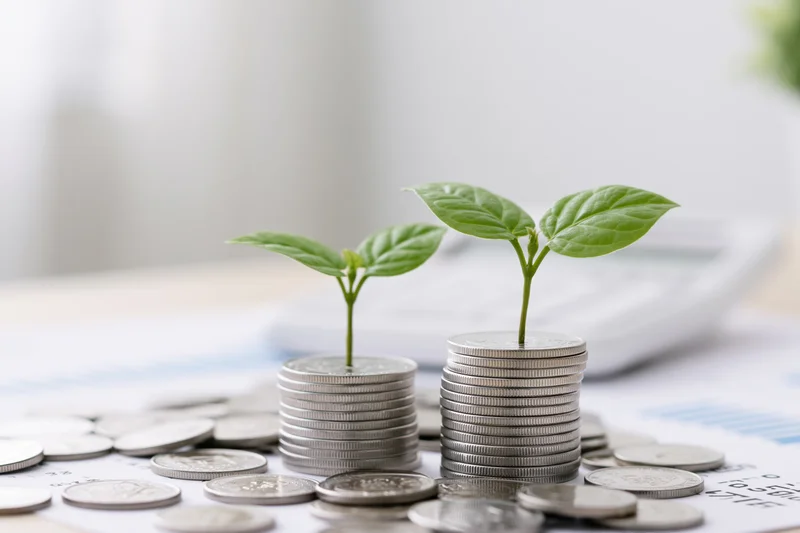 Two small green seedlings growing from stacks of silver coins on a financial document on a desk