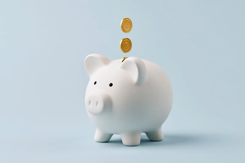 White ceramic piggy bank with gold coins falling into slot on light blue background