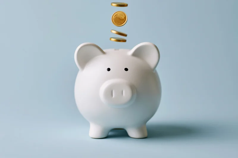 White ceramic piggy bank with gold coins falling into slot against light blue background