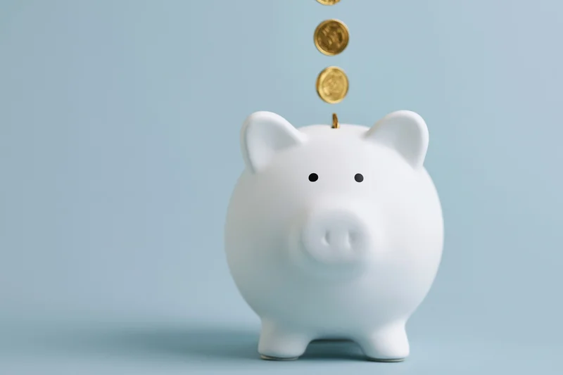White ceramic piggy bank with gold coins falling into slot against light blue background