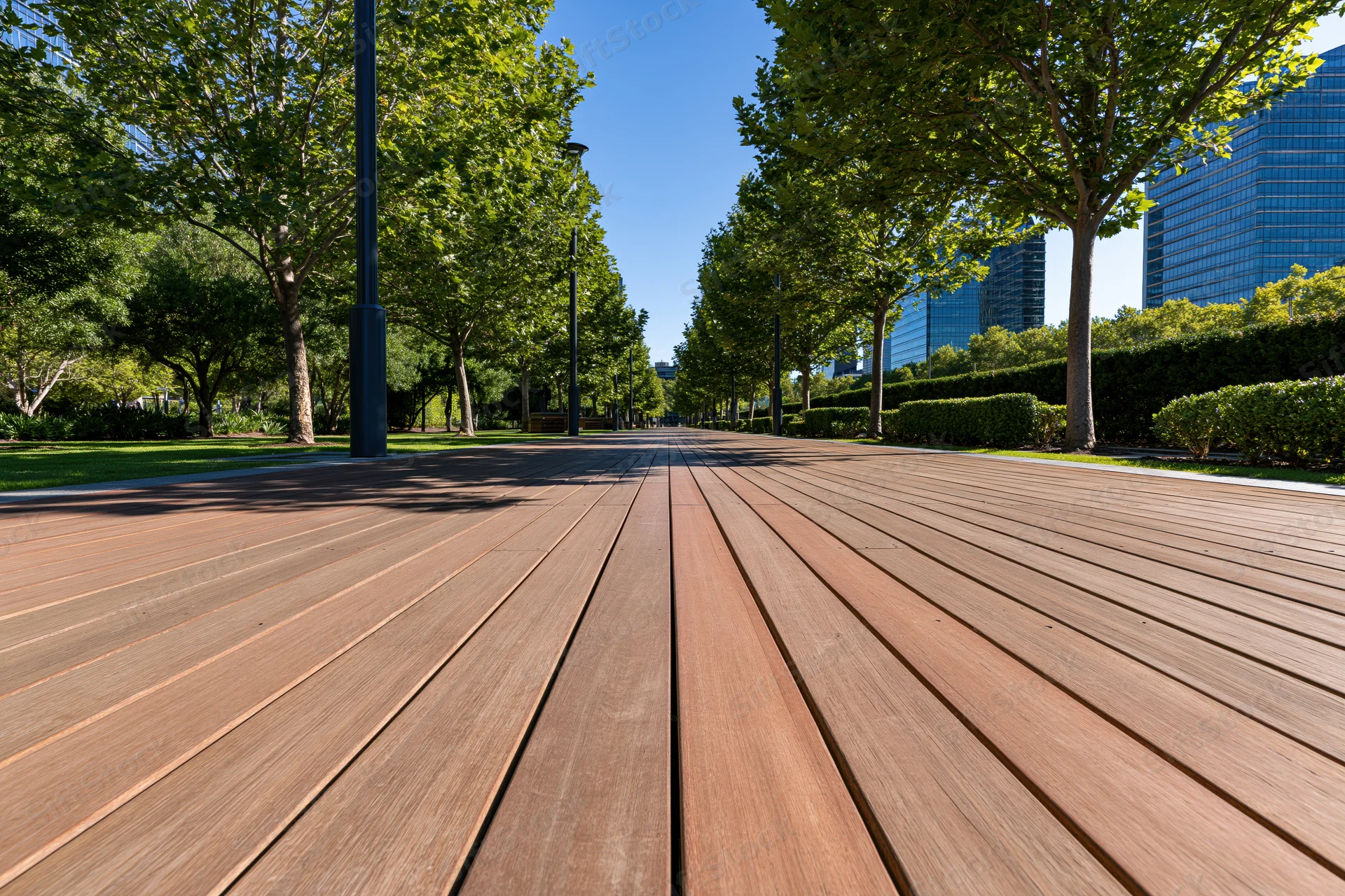 Wooden boardwalk path receding into the distance through a tree-lined urban park with skyscrapers in the background