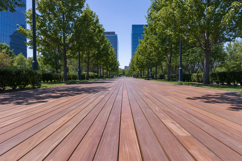 Wooden boardwalk path lined with trees and lampposts leading toward modern skyscrapers in an urban park