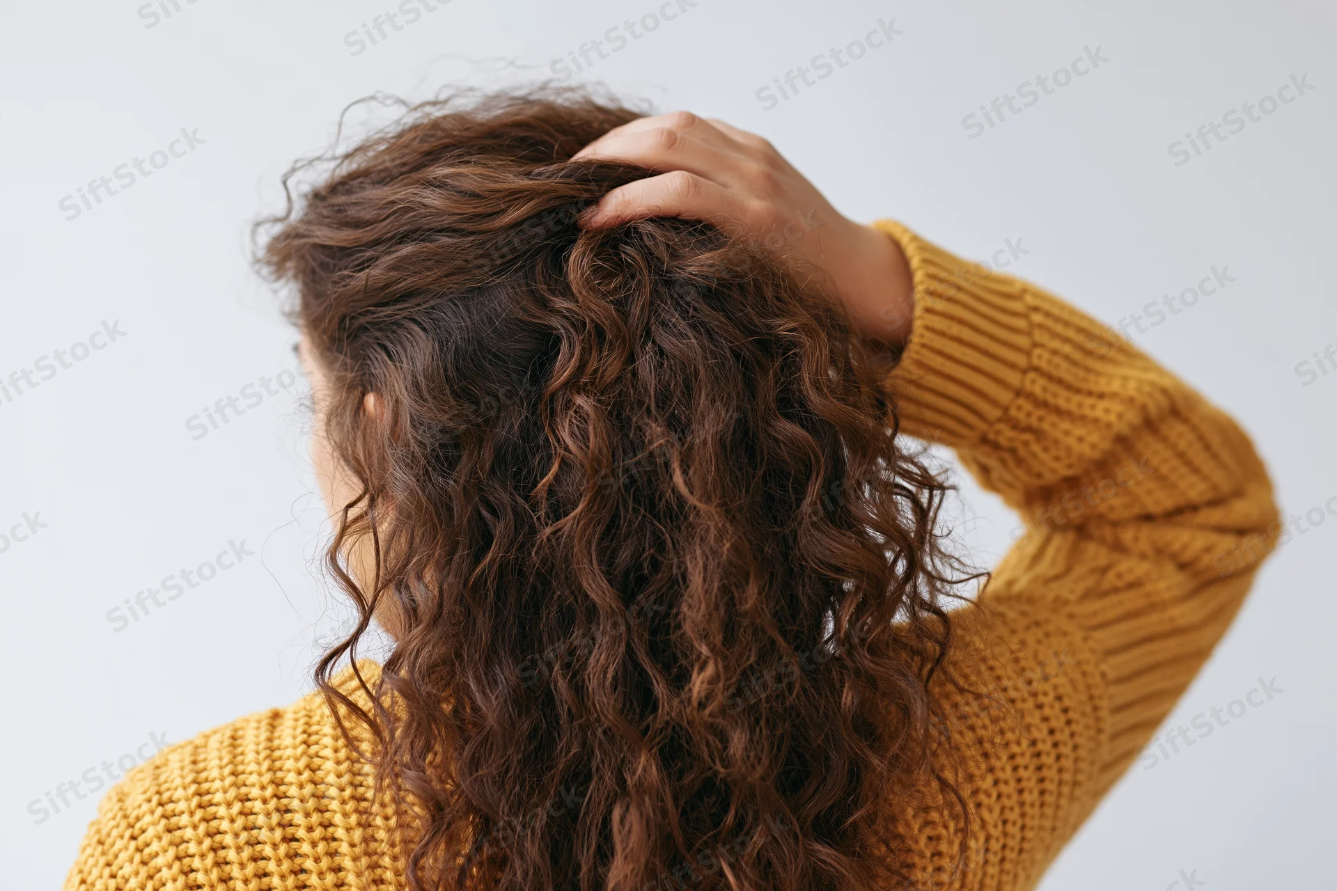 Woman from behind touching her long curly hair while wearing a yellow knit sweater
