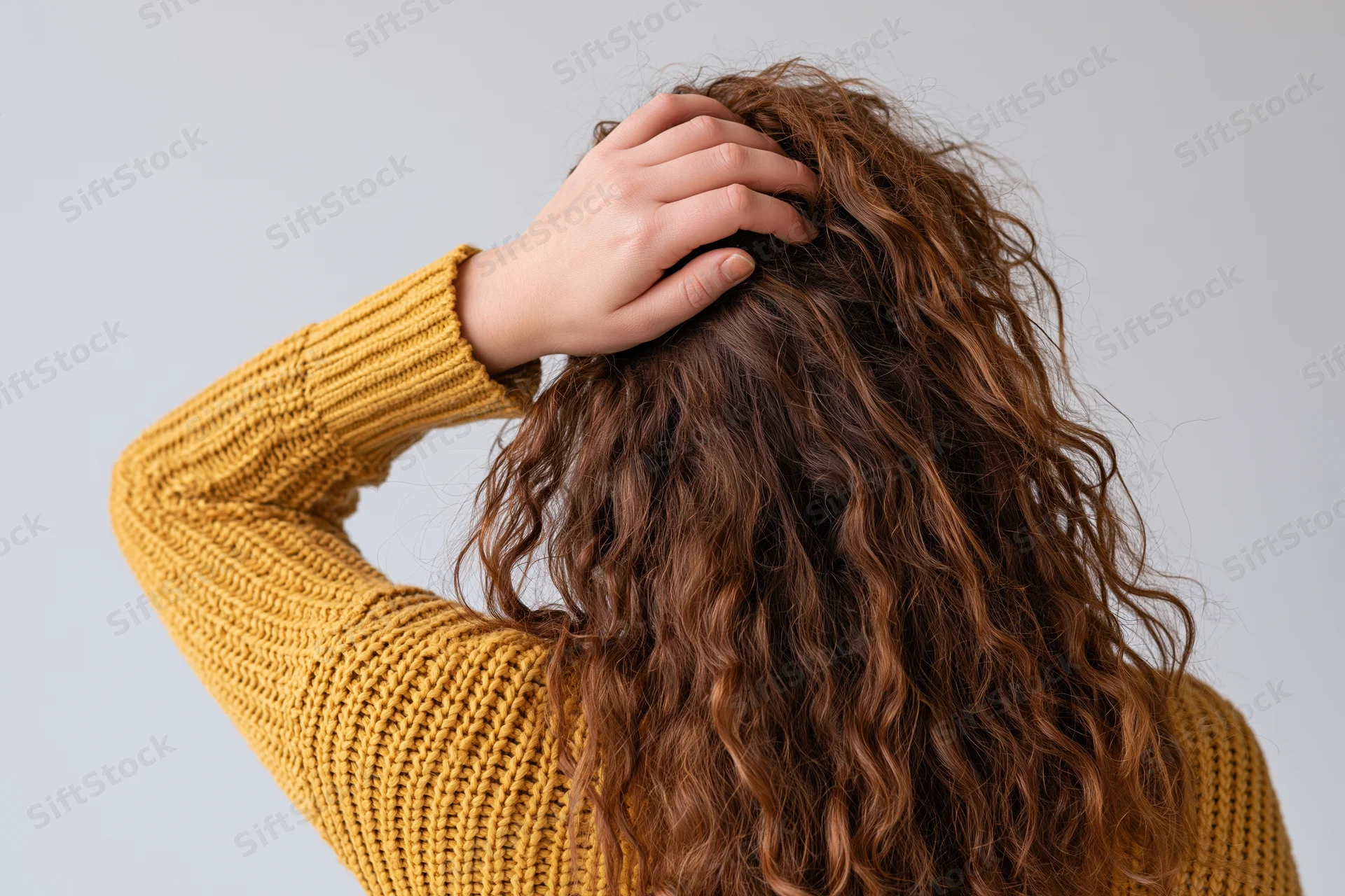 Woman from behind touching her long curly frizzy hair with hand, wearing yellow knit sweater