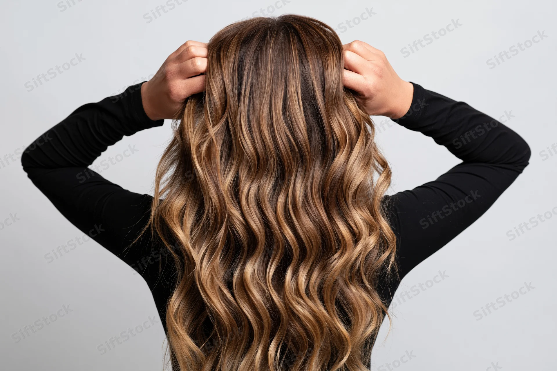 Woman with long wavy brown and blonde hair seen from behind, hands raised to head