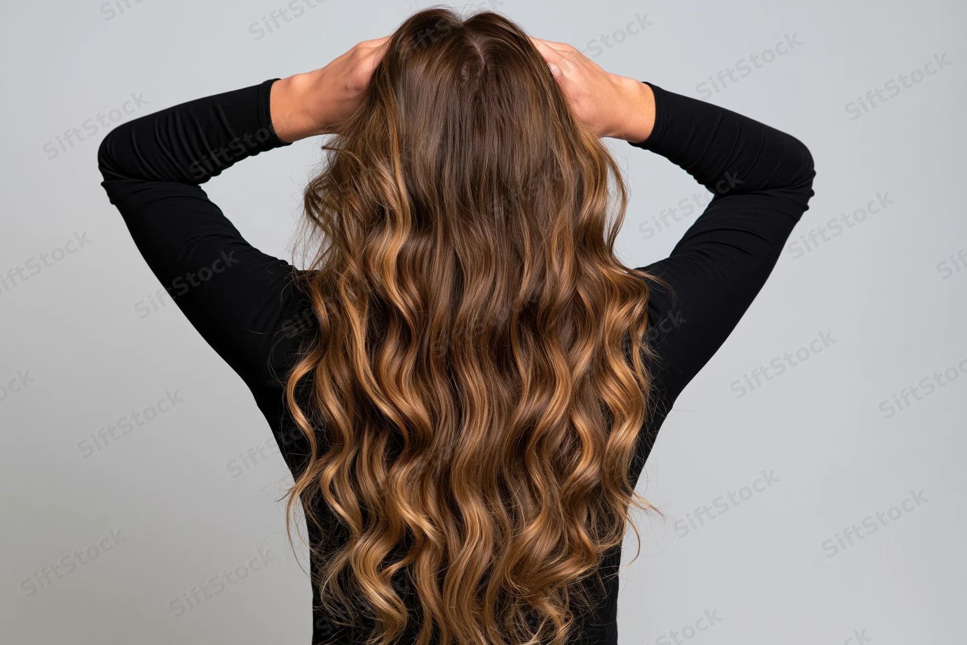 Woman from behind with hands raised to her long wavy hair in studio setting
