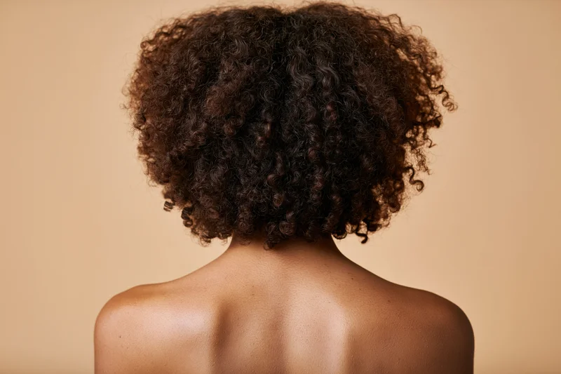 Back view of a black woman with natural curly hair showing bare shoulders against a neutral background