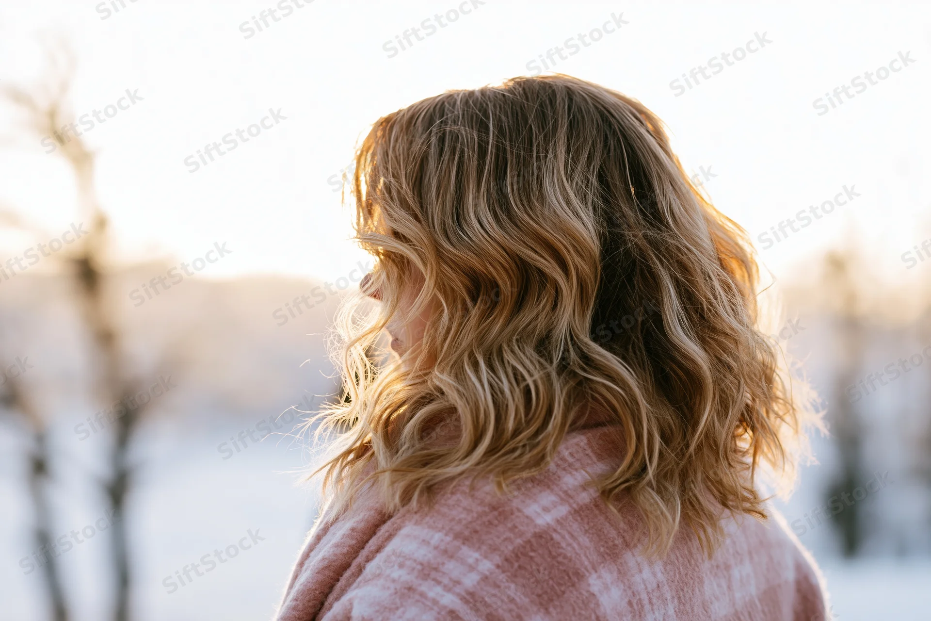 Woman with wavy blonde hair seen from behind in snowy landscape at sunset