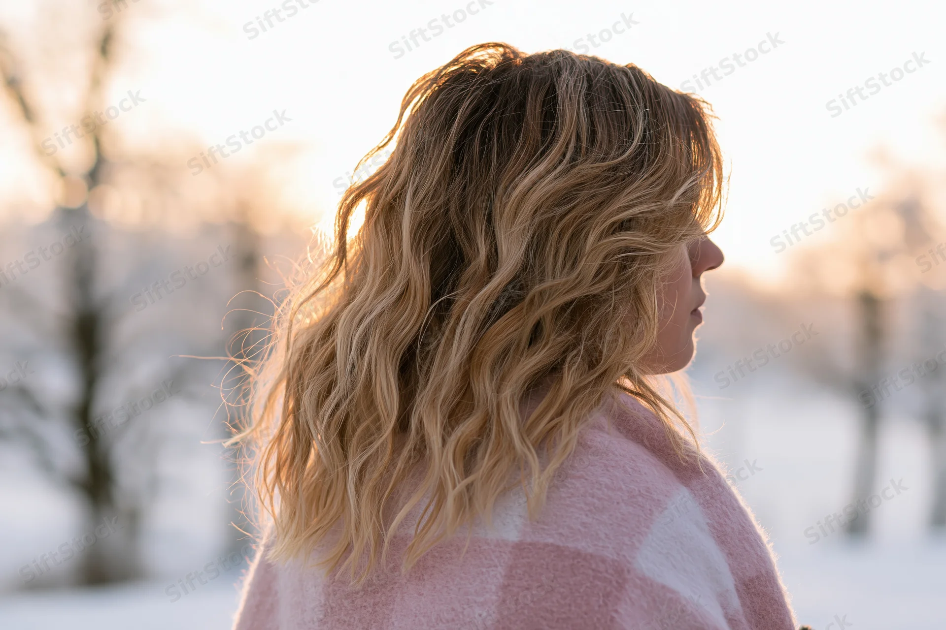 Young woman with wavy blonde hair in profile wearing pink checkered coat outdoors in snowy winter landscape at golden hour