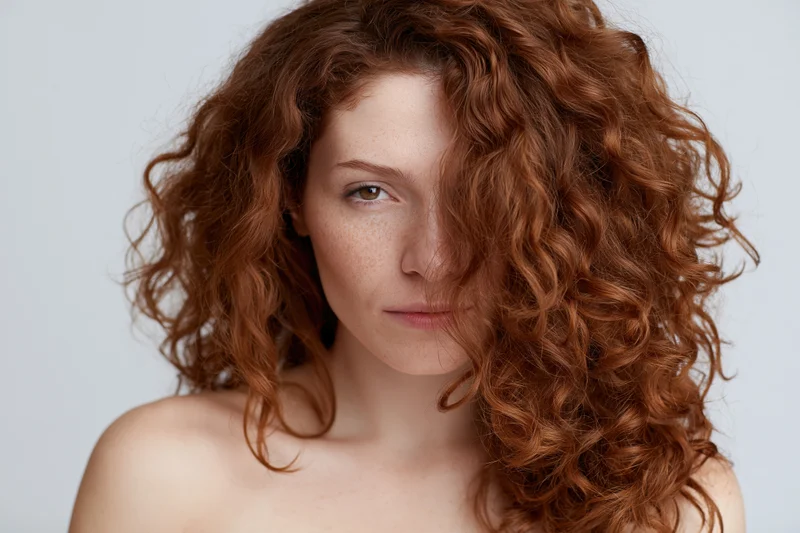 Young woman with voluminous auburn curly hair partially covering her face in studio portrait