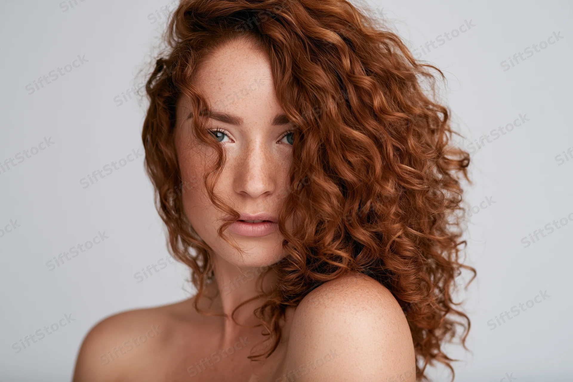 Young woman with voluminous auburn curly hair partially covering her face in studio portrait