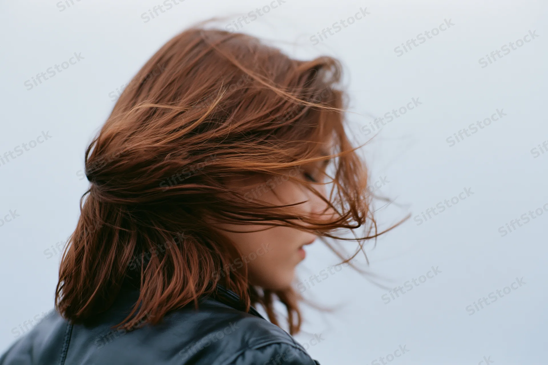 Woman with auburn hair blowing across her face in wind, wearing leather jacket outdoors