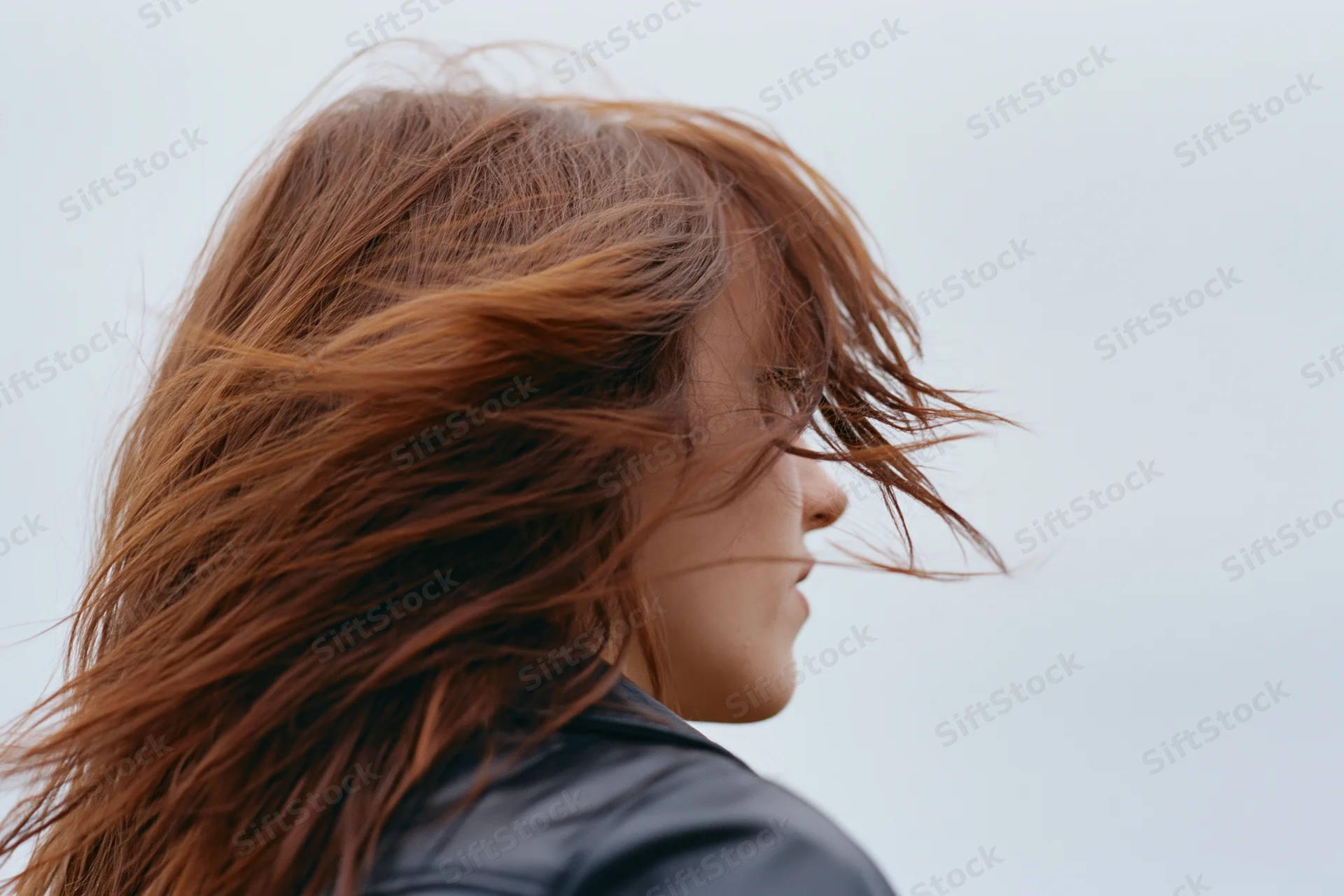 Young woman in profile with auburn hair blowing across her face in the wind outdoors
