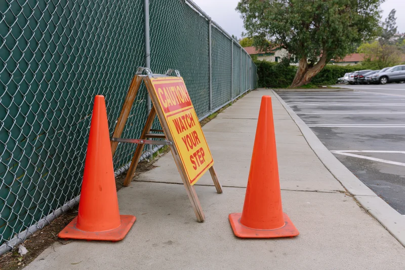 Orange traffic cones and a-frame caution sign on sidewalk beside chainlink fence near parking lot