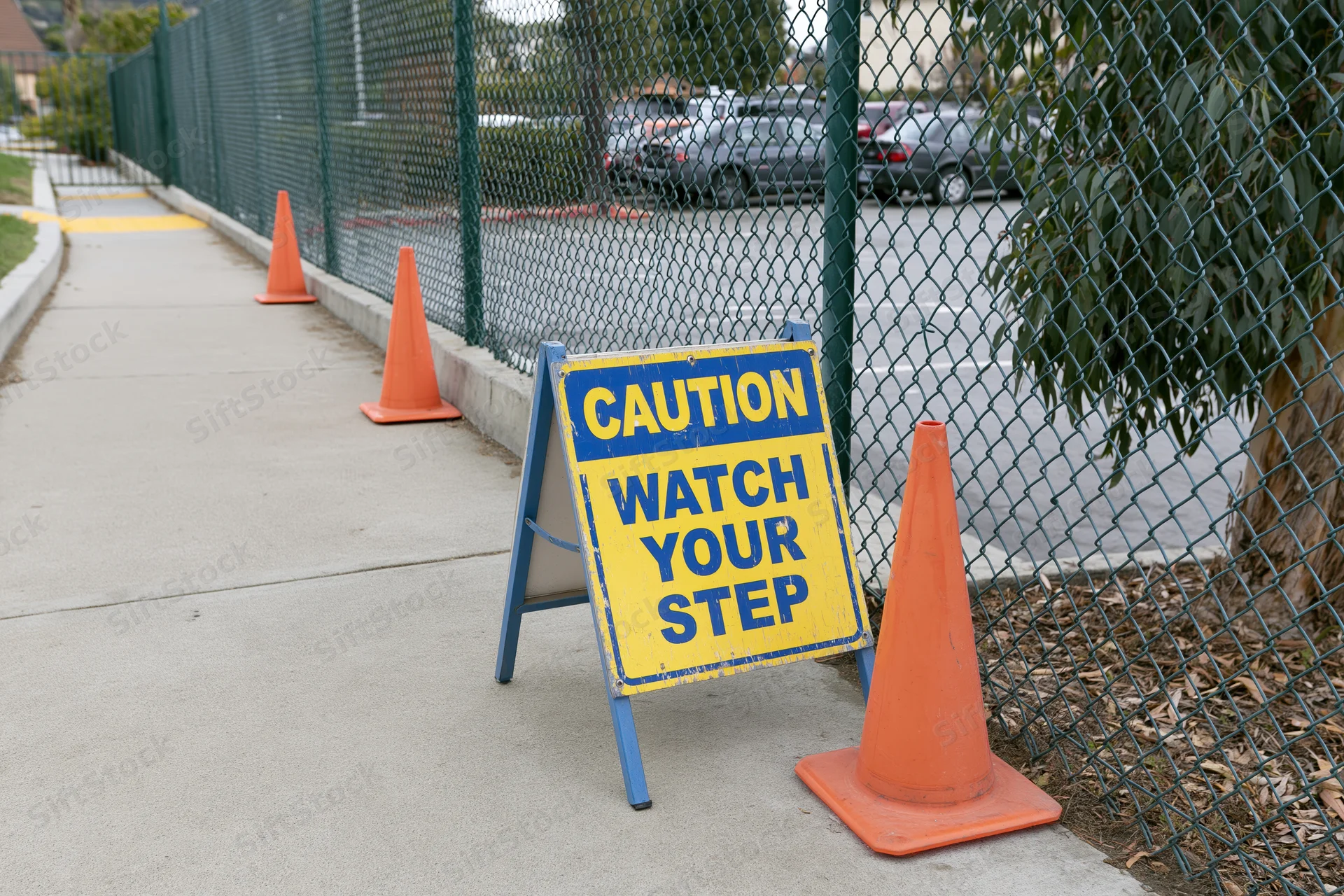 Caution sign reading watch your step on sidewalk flanked by orange traffic cones near chainlink fence