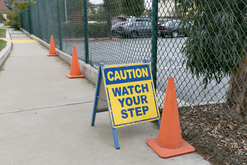 Caution sign reading watch your step on sidewalk flanked by orange traffic cones near chainlink fence