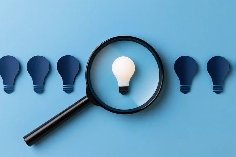 Magnifying glass focusing on a glowing lightbulb among dark ones on a blue surface