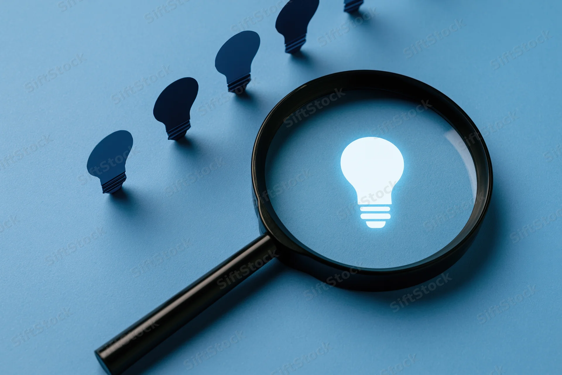 Magnifying glass focusing on a glowing lightbulb among dark silhouettes on blue paper