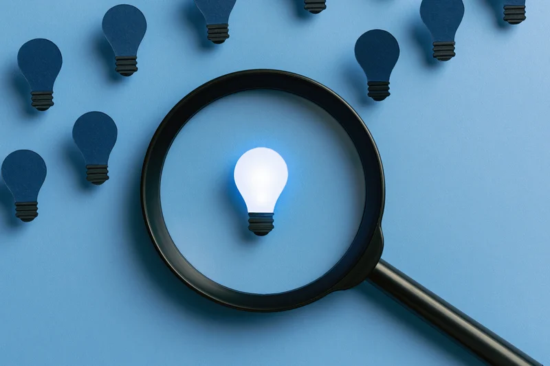 Magnifying glass over a glowing lightbulb on blue paper surface