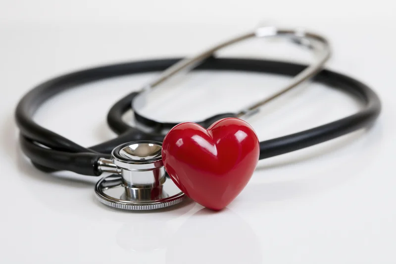 Black stethoscope coiled around a small red heart figurine on white surface